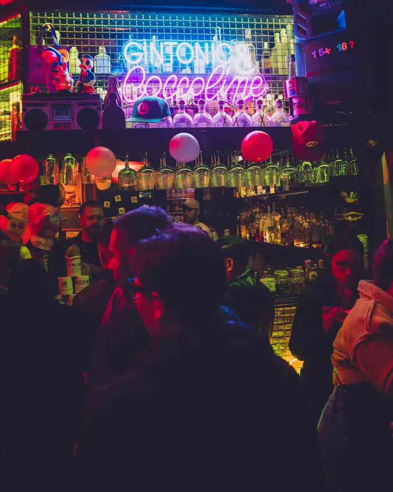 Il bancone del 7COINS sotto il neon «Gintonic & Coccoline», con la bottigliera, i bicchieri appesi e la gente al banco