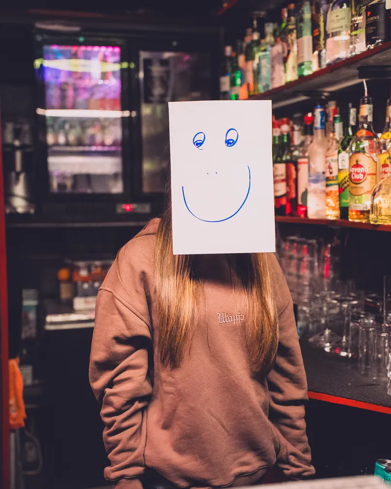Una barista al bancone con un foglio disegnato a faccina sorridente davanti al viso