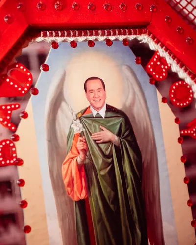 Un santino ironico con le ali d'angelo, in una cornice rossa piena di lucine