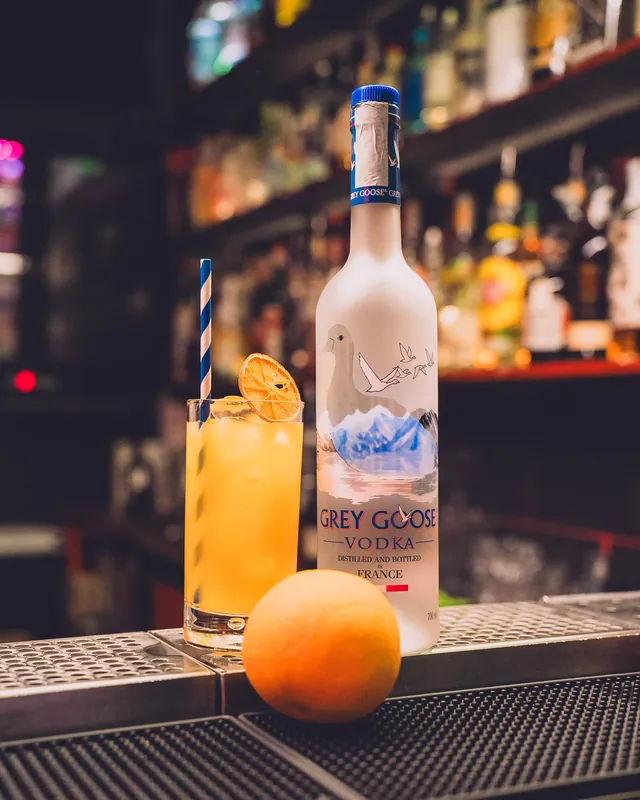 Long drink all'arancia con una fetta d'arancia essiccata, accanto a una bottiglia di vodka Grey Goose sul bancone
