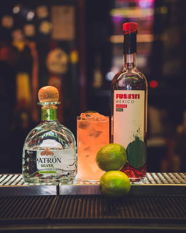 Una bottiglia di tequila Patrón Silver accanto a un drink con ghiaccio, due lime e una bottiglia di bitter