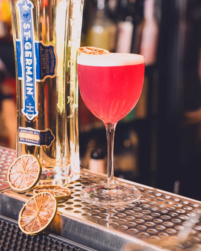Cocktail rosa con schiuma in una coppa, accanto a una bottiglia di St-Germain e a fette d'arancia essiccate